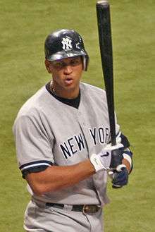 A man in a baseball jersey and batting helmet. His helmet is emblazoned with a white "N" and "Y" intertwined, and "NEW YORK" is stitched in black letters across his button-down jersey. The player is holding a black baseball bat almost vertically with black, gray, and white gloves.