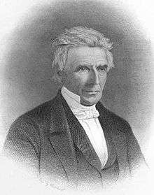 A man in his mid-60s turned a quarter of the way to the right. He's wearing a dark formal suit with a white shirt, high collar and white tie. His hair is white and loosely combed. His expression appears serious but not unfriendly.