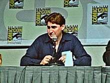 An image of a man in his 50s sitting at a green table and speaking into a microphone. He has brown hair and is wearing a blue shirt. in the background, a wall with the words "comic con" written on it can be seen.