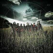 A group of nine men wearing masks stand in a field of grass with a gloomy clouded sky behind them.