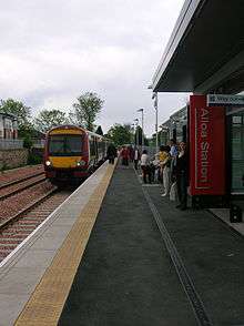 A railway platform with a train stopped at the station. A few passengers are scattered along the platform