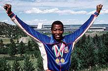 An African-American athlete wearing a blue running top and two gold medals hang from his neck.