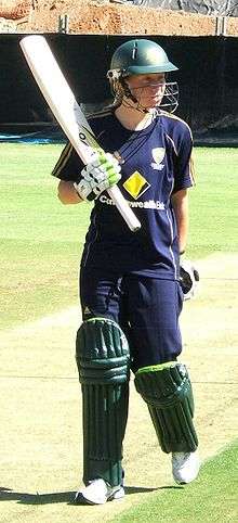 Young woman with a short blonde ponytail wearing a dark blue T-shirt, baseball cap and trackpants with gold stripes. Advertising logos of Adidas and Commonwealth Bank are present on the clothes. She is wearing a green helmet with a grill and pads on her legs, gloves and is holding a bat in her right hand.