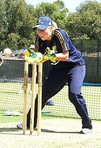 Young woman with a short blonde ponytail wearing a dark blue T-shirt, baseball cap and trackpants with gold stripes. Advertising logos of Adidas and Commonwealth Bank are present on the clothes. She is wearing gloves, standing with bent knees and open hands next to cricket stumps watching carefully for a white ball to catch.