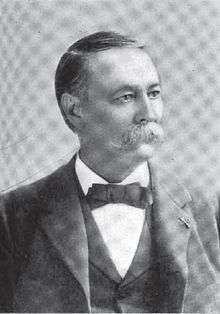 A white man with a full mustache in a dark suit with a bow tie and vest, looking right.