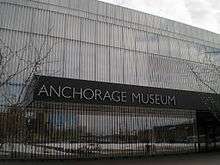 The mirrored facade of a building is seen with a stainless steel nameplate reading Anchorage Museum.