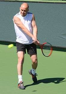 An older, bald man plays tennis.  He's wearing a white sleeveless shirt and black shorts.  He is bald and is holding a red tennis racket.