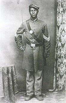 A young black man wearing a tilted forage cap and a long military jacket with a belt, a wide leather strap across the chest, and three chevrons on each sleeve. His right hand is holding a sword vertically.