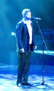 Johnston, a thick-set, teenage boy with short, spiked hair stands on stage and sings into a microphone. He is wearing a dark blazer, dark trousers and black shoes, with a white shirt and no tie.