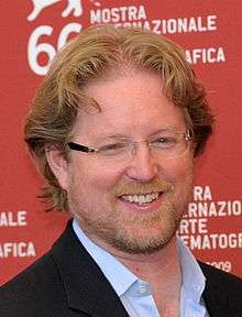 A photo of a middle aged man with red hair. He is wearing glasses, a white collared shirt, and a black coat.