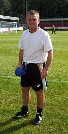 A blond-haired man in his 40s, wearing a white T-shirt and black shorts, stands near the edge of a football pitch.