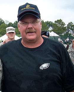 Color head-and-shoulders photograph of stocky white man (Andy Reid) wearing a dark green Philadelphia Eagles sweatshirt and a navy blue baseball cap with gold U.S. Marine Corps logo.