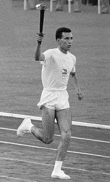 Athlete in white shorts and t-shirt running on an athletics track with the Olympic torch aloft in his right hand.