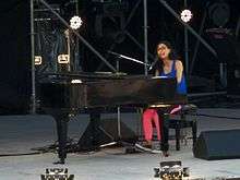 A woman with black glasses seated at a grand piano on a stage, wearing a blue singlet, pink tights and sneakers.