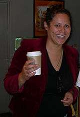 A smiling woman wearing a red jacket leaning on a bench holding a cup. Her black hair is pulled back.