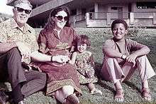 A young boy (preteen), a younger girl (toddler), a woman (about age thirty) and a man (in his mid-fifties) sit on a lawn wearing contemporary c.-1970 attire. The adults wear sunglasses and the boy wears sandals.