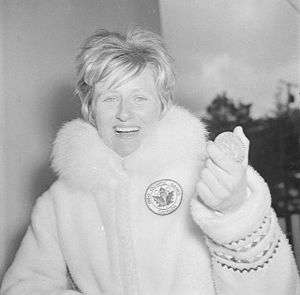 A woman with short blonde hair wearing a thick fur coat bearing a maple leaf and Olympic rings. She displays a large smile as she holds up an Olympic medal.
