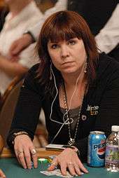A woman with shoulder-length brown hair wearing a black jacket and jewelry, with white earphones hanging from her ears to the iPod that sits on the poker table in front of her. On the table are poker chips, cards, a soda can, and a small bottle of water.
