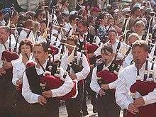 Group of young men and women, wearing white shirts (some with black waistcoats) and black trousers, marching in a parade, in the sunshine. Each is playing a bagpipe. The bag is a claret colour. The entire picture is full of people. Those not taking part in the parade are watching the procession.