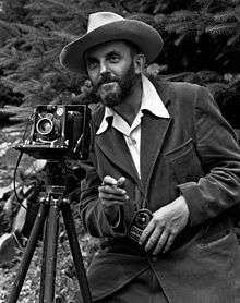 A photo of a bearded Ansel Adams with a camera on a tripod and a light meter in his hand.  Adams is wearing a dark jacket and a white shirt, and the open shirt collar is spread over the lapel of his jacket.  He is holding a cable release for the camera, and there is a rocky hillside behind him.  The photo was taken by J. Malcolm Greany, probably in 1947.