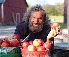 Photo of a white man with a gray beard and wild grey hair, sitting in front of a bushel of apples and holding an apple, and grinning broadly