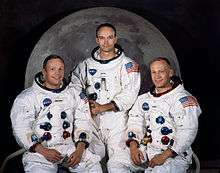 Three astronauts in spacesuits without helmets sitting in front of a large photo of the Moon.
