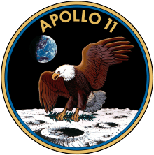Circular insignia: Eagle with wings outstretched holds olive branch on Moon with Earth in background, in blue and gold border.