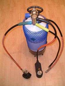 A large scuba cylinder is shown, with a handle, boot, plastic net and single hose regulator with one demand valve, a combo submersible pressure gauge console and two low-pressure inflator hoses.