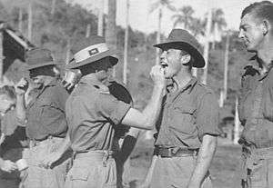 Men in Army uniforms with slouch hats stand in a row. One has his mouth open while another man in Army uniform with a slouch hat places something inside.