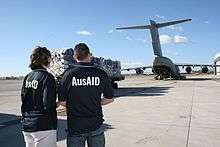 Colour photo of a woman and a man wearing blue polo shirts with "AusAID" on the back looking at a large military transport aircraft at an airport. A trailer loaded with supplies is in front of the two people.