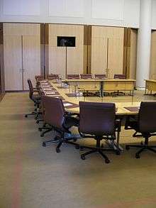 A meeting room containing a large horseshoe-shaped desk, with red leather office chairs surrounding its outside edge, a microphone mounted in the desk in front of each chair