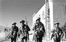 Soldiers wearing leather jerkins and helmets walk past whitewashed buildings with rifles slung