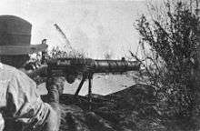 A soldier is looking through the sights of a machine gun amongst the grass in the prone position.