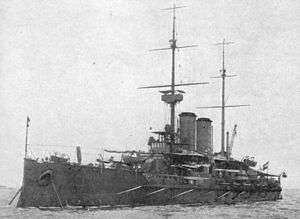 A large battleship at anchor in calm waters with two tall masts and two round funnels.