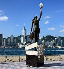A bronze statue on a pedestal, with the city skyline in the background. The pedestal is designed in the image of four clapperboards forming a box. The statue is of a woman wrapped in photographic film, looking straight up, with her left hand stretched upwards and holding a glass sphere containing a light.