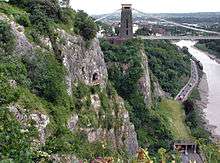 Rocky side to a gorge with a platform in front of a cave halfway up. To the right are a road and river. In the distance are a suspension bridge and buildings.