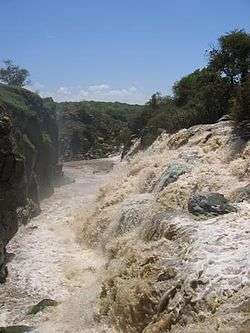 A picture of a waterfall to the right, shooting water into the side of a rapid flowing river.