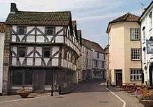 Street scene. On the left of the road is a half timbered house where the first and second storeys have irregular black wooden beams showing through white painted walls.