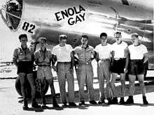 A Silver aircraft with "Enola Gay" and "82" painted on the nose. Seven men stand in front of it. Four are wearing shorts, four are wearing T-shirts, and the only ones with hats have baseball caps. Tibbets is distinctively wearing correct uniform.