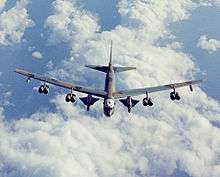 Aerial view of B-52 fly above white clouds and the sea. It carries two triangular-shaped vehicles under the wings between the fuselage and inboard engines.