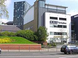 A quirky modern five-storey building with a large sign saying "BBC Yorkshire" in black above the second-floor windows on the white-fronted façade of the lower four floors can be seen on the far side of a dual-carriageway road with a barrier along the central reservation. At right-angles to the right of the building is a tall blue slab with the letters "BBC" in white at the top. The left side of the building is mostly brick-red with a few windows, but above it is a light blue windowless section. The roof above this and the grey fifth floor of the frontage curves gently down to the rear. A lone car is driving from left to right along the road; between it and the building, temporary boards have been erected in front of a building to the left. In the top left-hand corner of the picture, part of a tall many-windowed building can be seen.