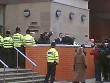 Two suited men wave from behind a red brick wall, at the top of a short flight of steps leading to a grey building. Several police officers are in attendance.