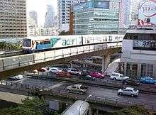 An elevated train, painted in blue, white and a red stripe and with advertisements with the name "acer", running above a road lined with many tall buildings and crossing an intersection with a flyover bridge with many cars