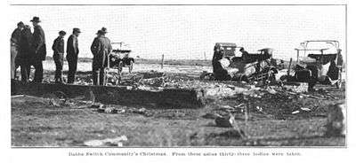 Men in hats and coats talk in a field full of debris, including a segment of a brick foundation to the left and burnt cars to the right.