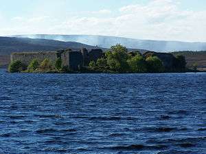  In the foreground are the blue waters of a lake. Beyond that is a wooded shoreline on which there are the ruins of a large walled structure. Smoke drifts across the moorland in the distance.