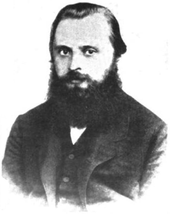 A man in his late 20s or early 30s with dark hair and a bushy beard, wearing a dark coat, dress shirt and tie.