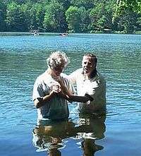 A baptism in a river. Two men are standing hip-deep in blue water with trees in the background.