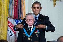 Head and torso of elderly white man in military dress uniform with the President standing behind him, placing a medal with a blue ribbon around his neck