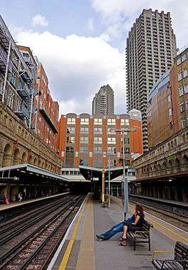 A picture of a railway station in a city taken from an elevated concrete platform, sheltered in the background, with two sets of two tracks each and sheltered platforms on either side. A man and a woman are sitting on a wooden bench in the foreground facing left. In the background the tracks disappear into two tunnel portals. Brick buildings rise on all three sides of the cutting; two large concrete towers rise behind them in the centre and on the right.