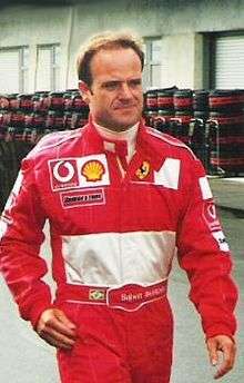 Head, arms and torso of a man in his thirties with his right arm slightly bending. He is wearing a scarlet red racing overalls which displays the Vodaphone, Shell, Bridgestone and Ferrari logos at the front with a white background embroiled on the bottom top.